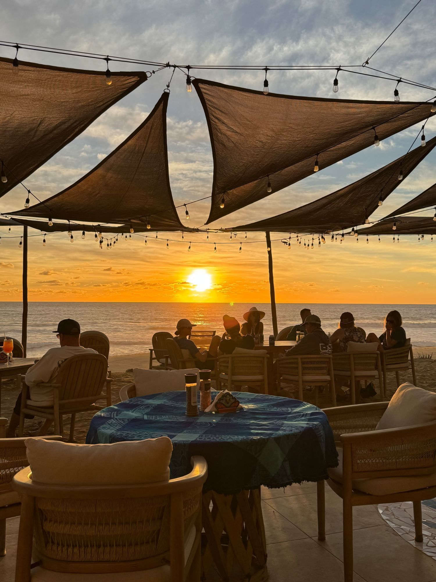 Guests at the tables of The Sunset Restaurant under shade sails and string lights, the sun dropping into the Pacific beyond